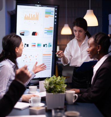 Multi-ethnic businesspeople discussing financial company solution sitting at conference table in meeting room. Diverse teamwork overworking at management strategy using tv late at night.