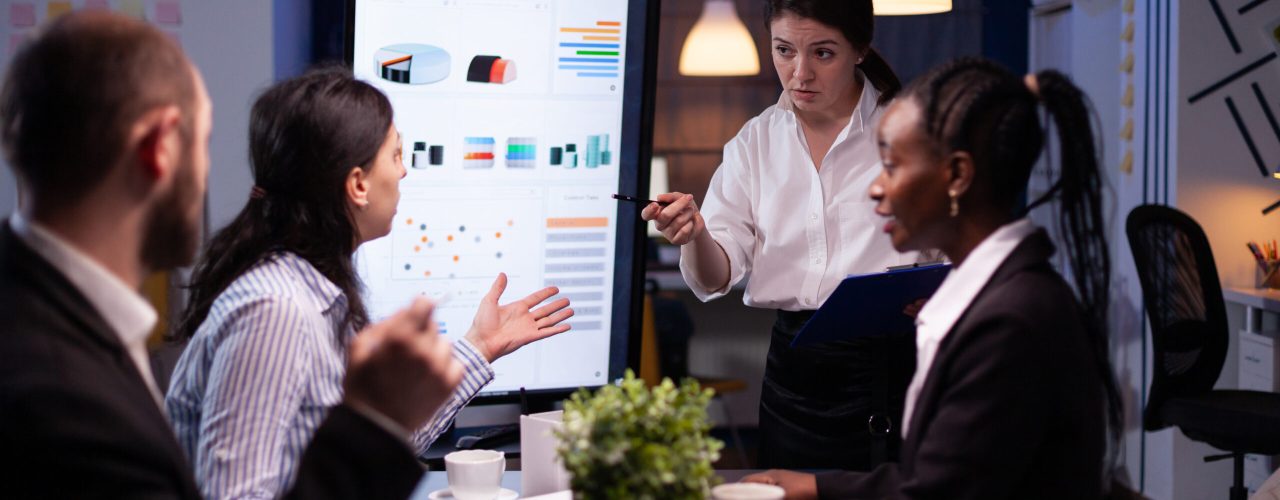 Multi-ethnic businesspeople discussing financial company solution sitting at conference table in meeting room. Diverse teamwork overworking at management strategy using tv late at night.