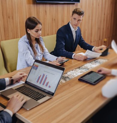 office workers like a team hold a meeting at one desk for laptops, tablets and papers, on the background a large TV set on a wooden wall