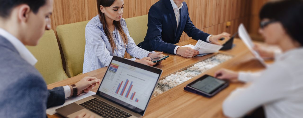 office workers like a team hold a meeting at one desk for laptops, tablets and papers, on the background a large TV set on a wooden wall