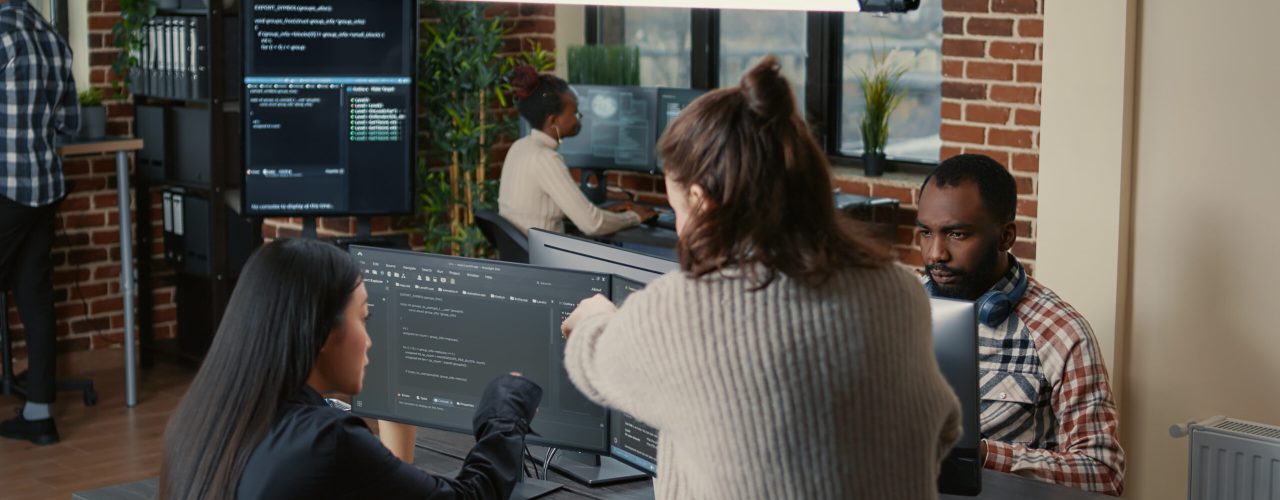 Software programer pointing pencil at source code on computer screen explaining algorithm to coworker standing next to desk. Programmers discussing online cloud computing with team.